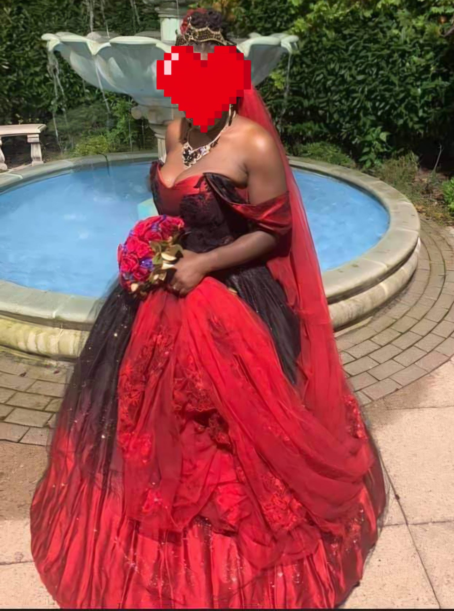 A woman wearing a voluminous off-the-shoulder ball gown in red and black, standing outdoors in front of a stone fountain. Her fave is covered with a pixelated heart 